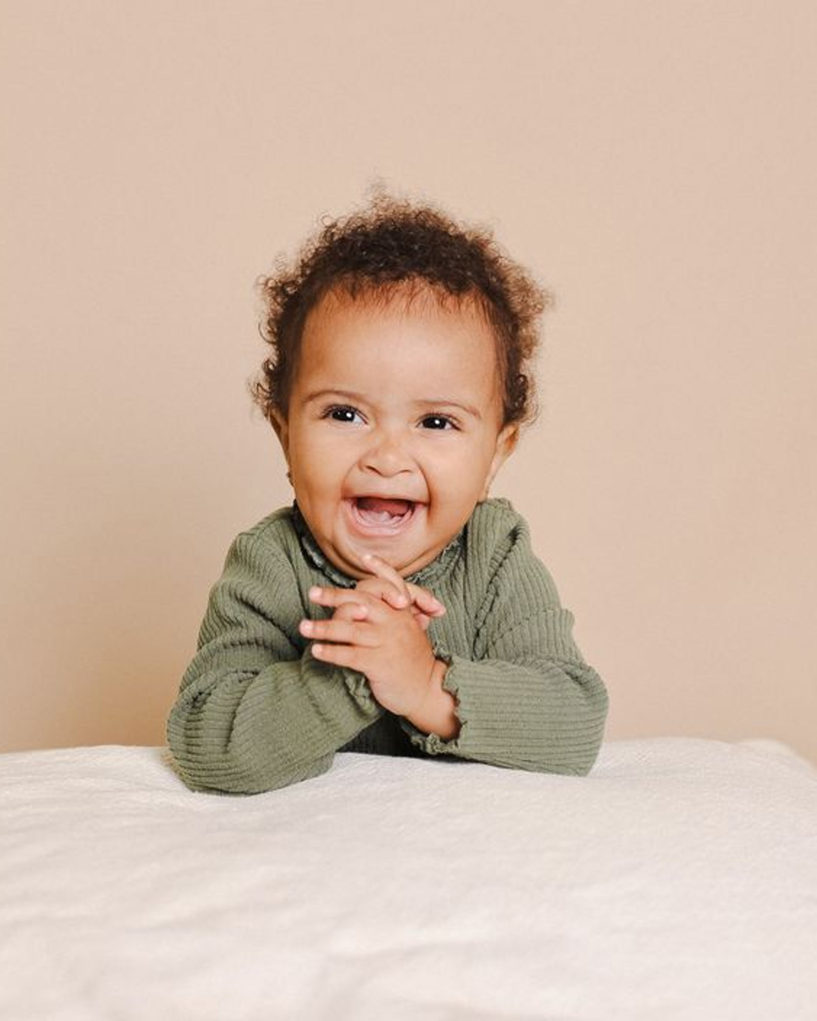 Studio portrait of Dixie laughing in an olive sweater, hands clasped on a white seamless